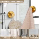 Two round brushed gold Fenas suction cup hooks mounted on a white tiled shower wall, holding a towel and a loofah.