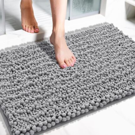 A soft, rectangular grey chenille bath mat with a shaggy texture, placed on a clean bathroom floor.